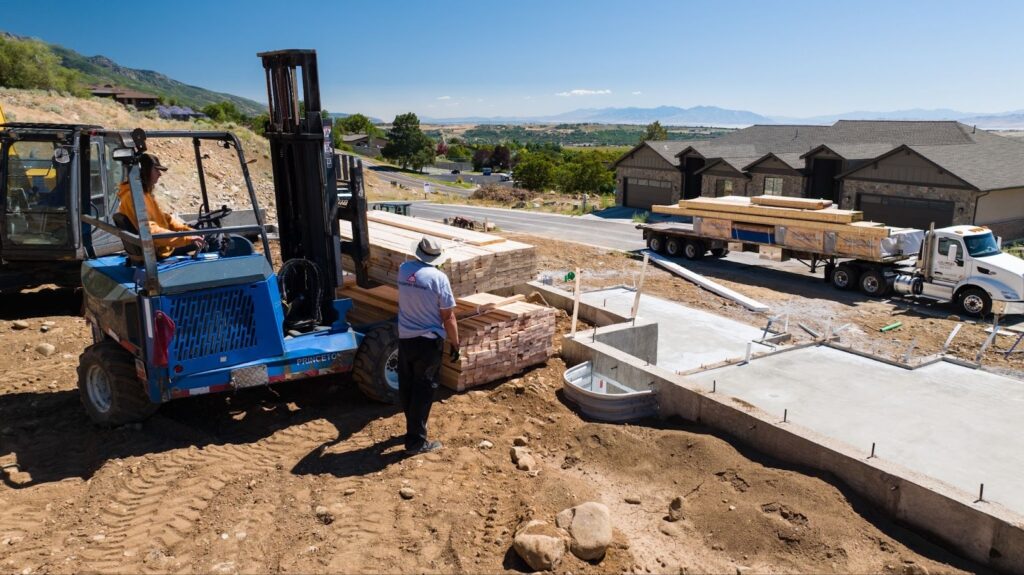 Forklift unloads bundled lumber at a residential construction site while a flatbed truck delivers materials near a concrete foundation in Utah.