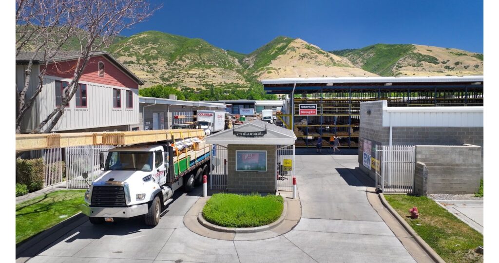 Colonial Building Supply yard entrance with a delivery truck loaded with lumber, with covered lumber racks visible and mountains in the background in Utah.